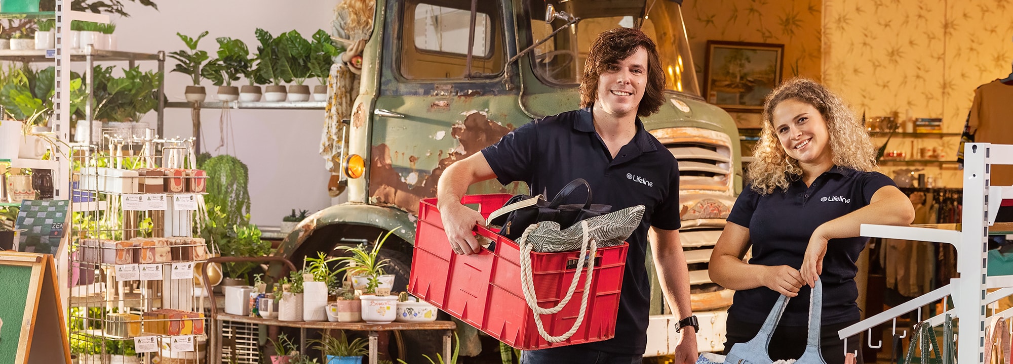 A young man and woman, wearing Lifeline T-shirts, stand in a Lifeline Shop surrounded by secondhand and new items, with a truck in the background.