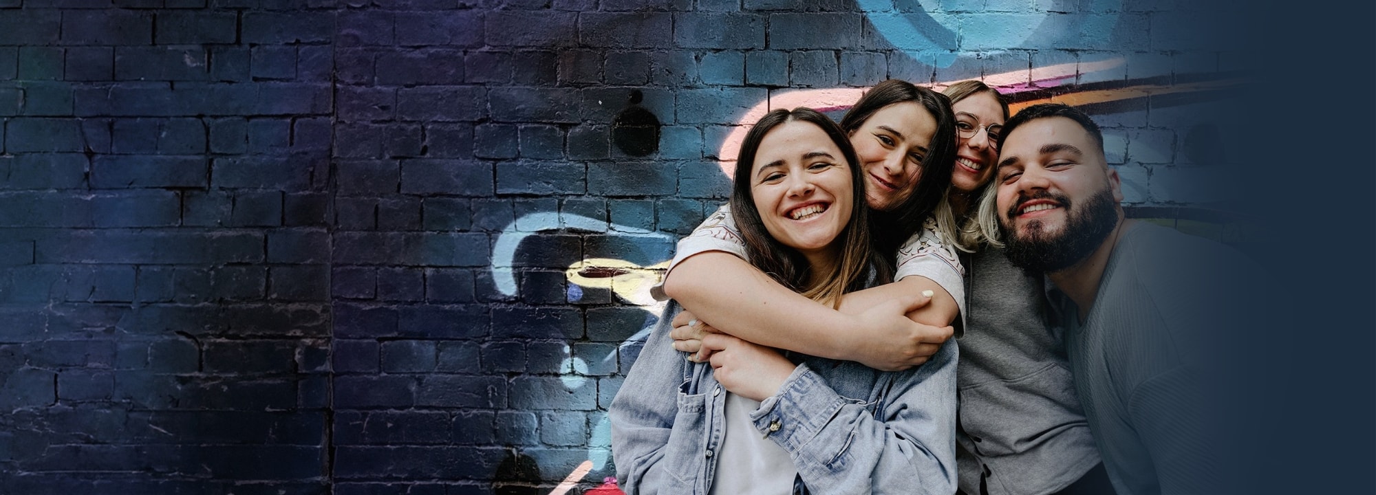 A group of friends are holding each other close and smiling warmly in front of a blue brick wall