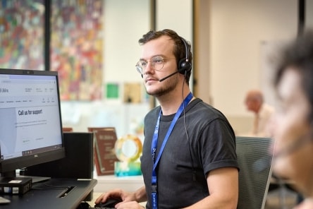 A young Crisis Supporter wearing glasses and a headset works at a computer, with a second Crisis Supporter in the foreground also wearing a headset.