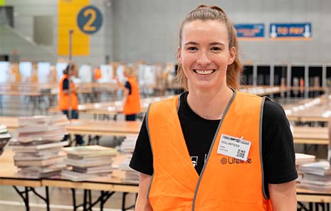 A feamle volunteer with gloves and hi-vis jacket in front of a table of books.