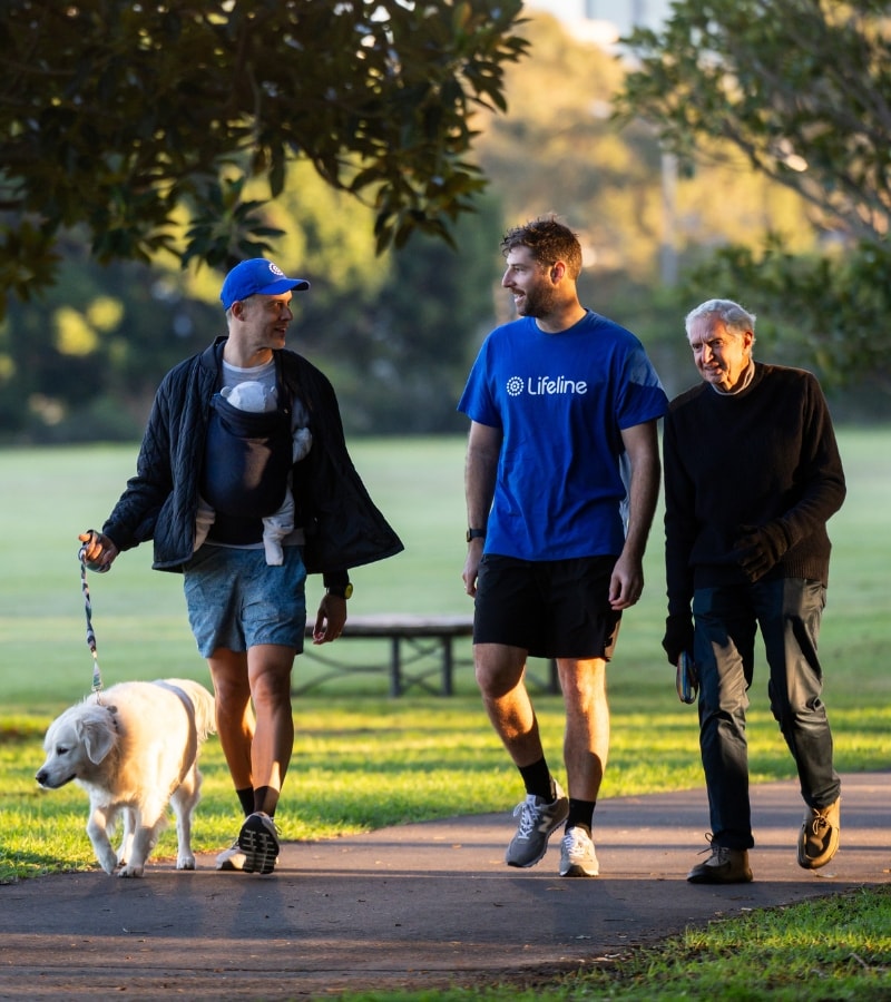 Three people and a dog walk along a path in a park at sunset. The person on the left is carrying a baby in a carrier and walking a dog on a leash. The person in the middle wears a blue t-shirt with the Lifeline logo.