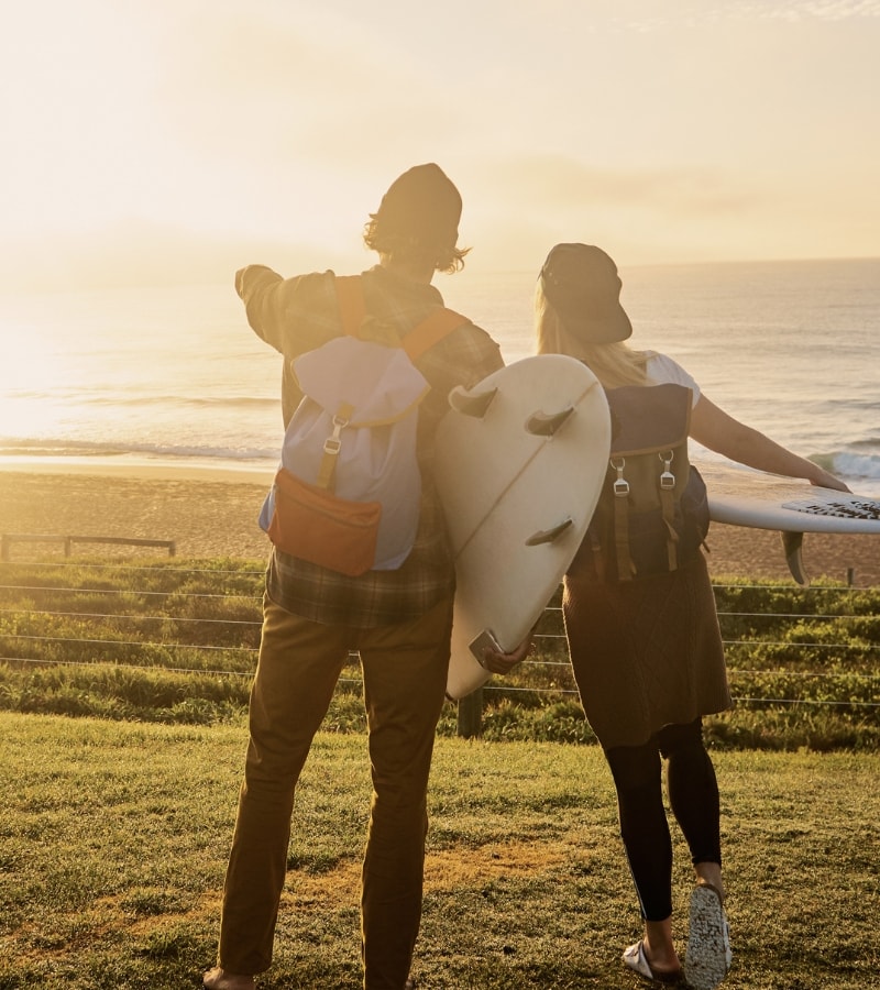 Two people with surfboards and backpacks stand on a grassy hill overlooking the ocean at sunrise.