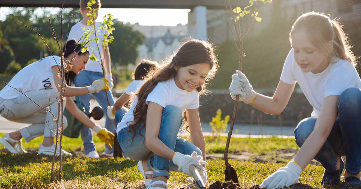 Two children plant a tree, as an adult in the background shows more children how to plant trees.