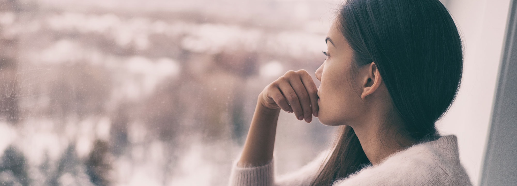 A young woman with long dark hair looks pensively out a rain-soaked window.