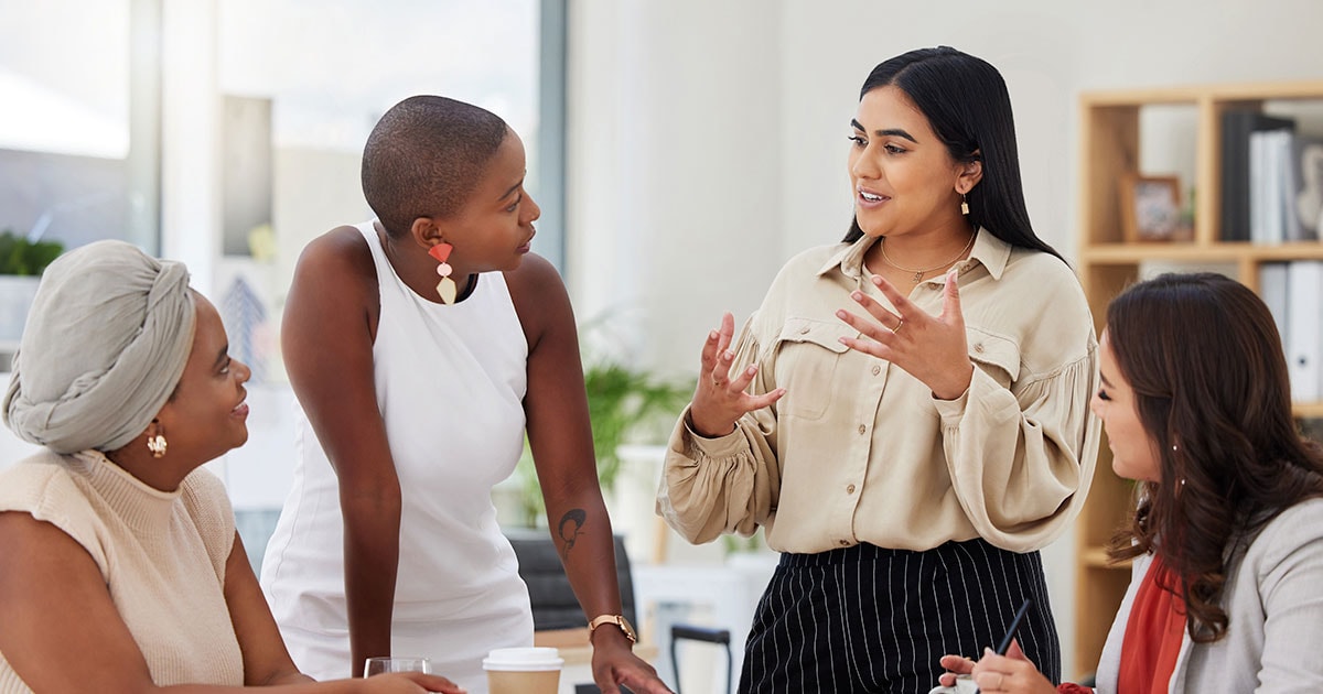Three young women gather around a work table listening to a fourth woman speaking animatedly.