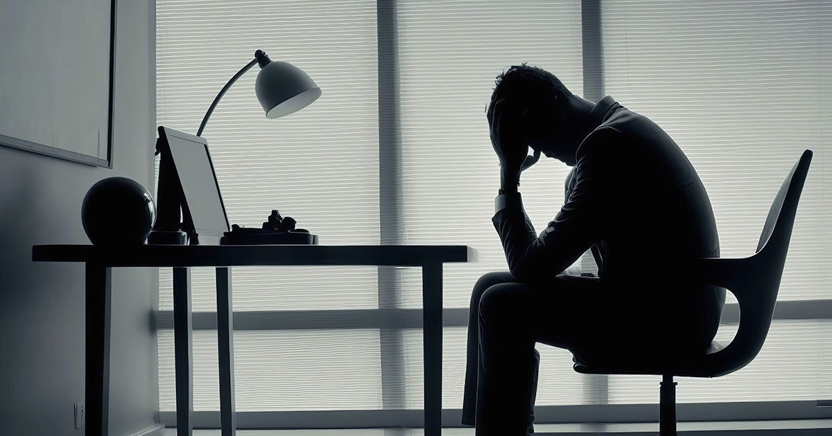 The silhouette of a man bowing his head in his hands as he sits at his desk in an office.