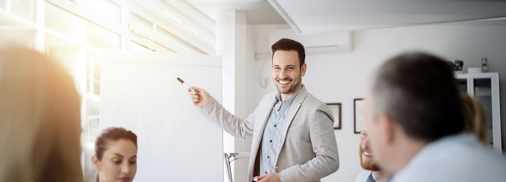 A smiling man in a suit jacket points at a blank whiteboard, as colleagues around a table watch.