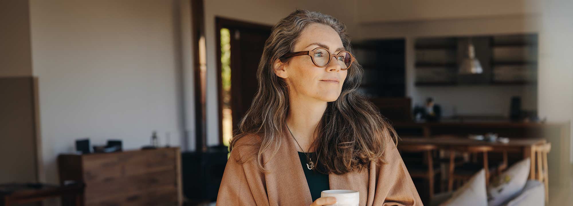 A woman with long brown hair and glasses, holding a cup in both hands in her loungeroom.