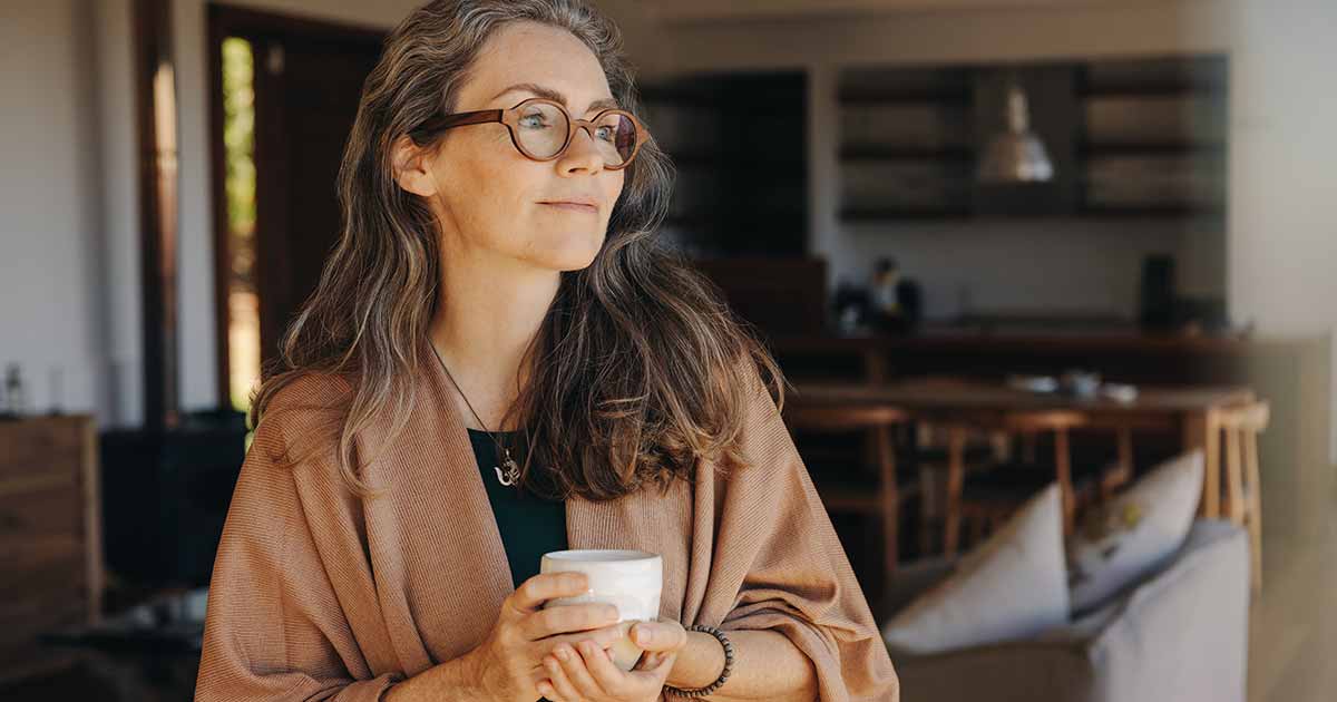 A woman with long brown hair and glasses, holding a cup in both hands in her loungeroom.
