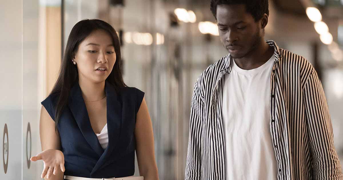 Two young people chat while walking through an office corridor.